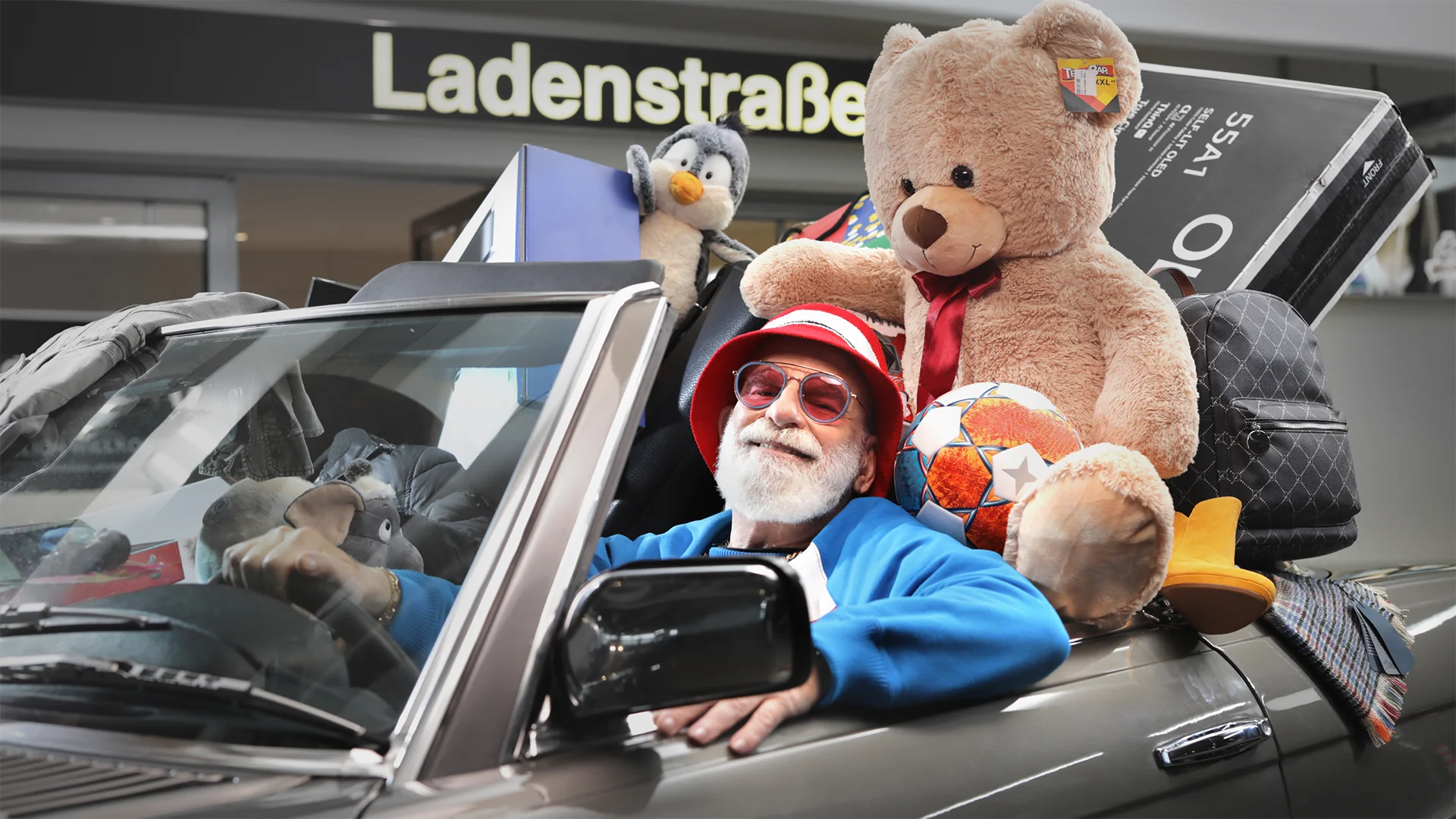 Ein Bild zeigt einen Mann im Cabriolet mit vielen Einkäufen zum Motiv 